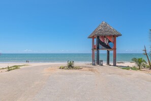 A beach pavilion with a thatched roof, a paved walkway, and a view of the ocean. - Eco Paradise #101 - Apt in Cumbuco by Carpediem (Caucaia)