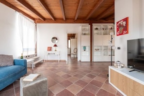 Ein Wohnzimmer mit einem blauen Sofa, einem Couchtisch und einem Fernseher auf einer Holzkommode. Eine weiße Schrankwand mit Regalen und ein Spiegel sind an der Wand angebracht.