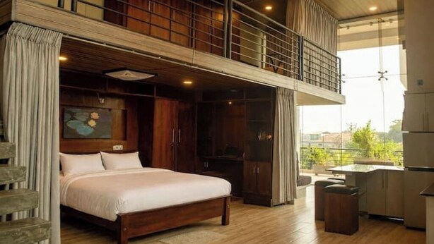A spacious bedroom with a large bed, wooden flooring, a balcony, and a view of the city.