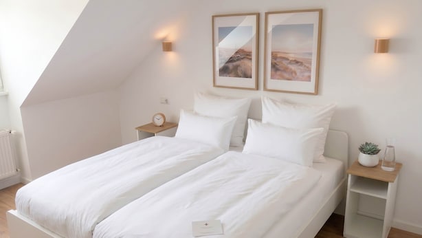 Un dormitorio con una cama doble, una mesita de noche con un reloj, un pequeño estante con una planta y dos cuadros en la pared.