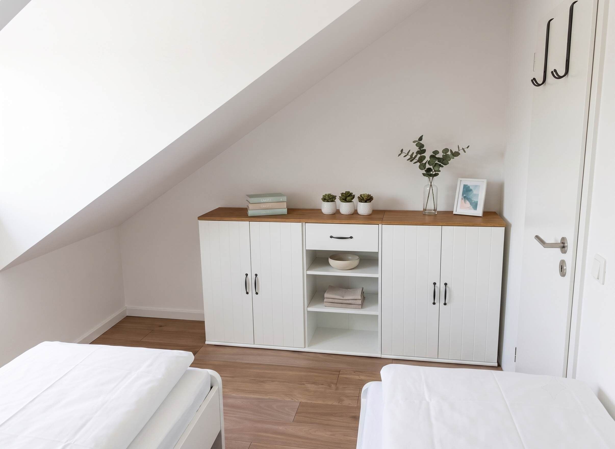 Ein Schlafzimmer mit schrägen Wänden, einem weißen Kleiderschrank, zwei Betten und einem kleinen Tisch mit Pflanzen.