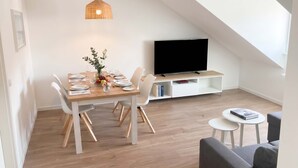 Un comedor con una mesa dispuesta para cuatro, un televisor sobre un mueble bajo, y una pequeña mesa redonda con un libro.