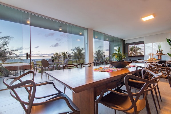 A spacious dining area with a large wooden table and modern chairs, large windows offering a view of palm trees and the ocean, and a cozy seating area with a small table and chairs.