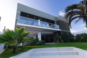 Un edificio moderno de dos plantas con techo plano, un balcón con barandilla de vidrio y un jardín cuidado con palmeras.