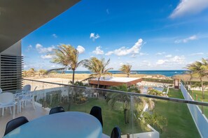 Un balcón con barandilla de vidrio que da a una playa, palmeras y una zona de piscina.