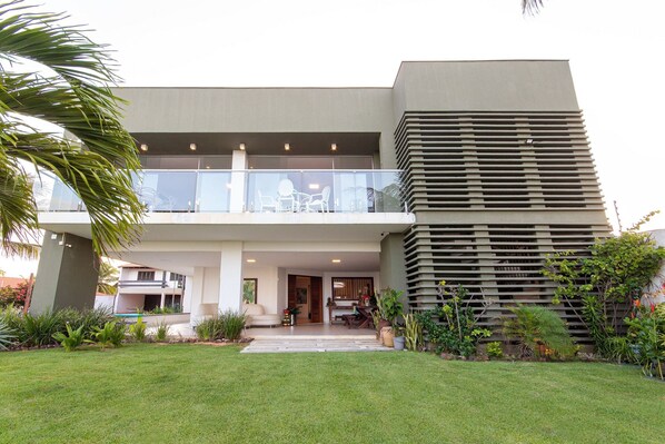 Un edificio moderno de dos pisos con techo plano, ventanas amplias y un balcón con mobiliario exterior.