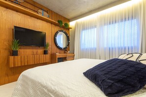 Un dormitorio con cama, televisión montada en la pared, escritorio de madera y ventana con cortinas.