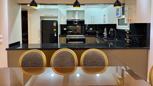 A modern kitchen with a black countertop, white cabinets, and three gold-accented chairs around a glass table. - Royal Gameat Al Dewal Residence (Giza Governorate)