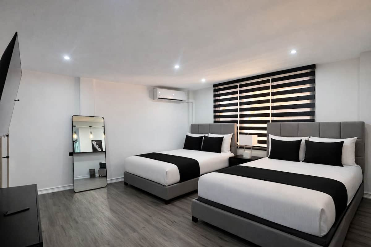 A modern hotel room with two beds, a mirror, a desk, and a window with blinds.