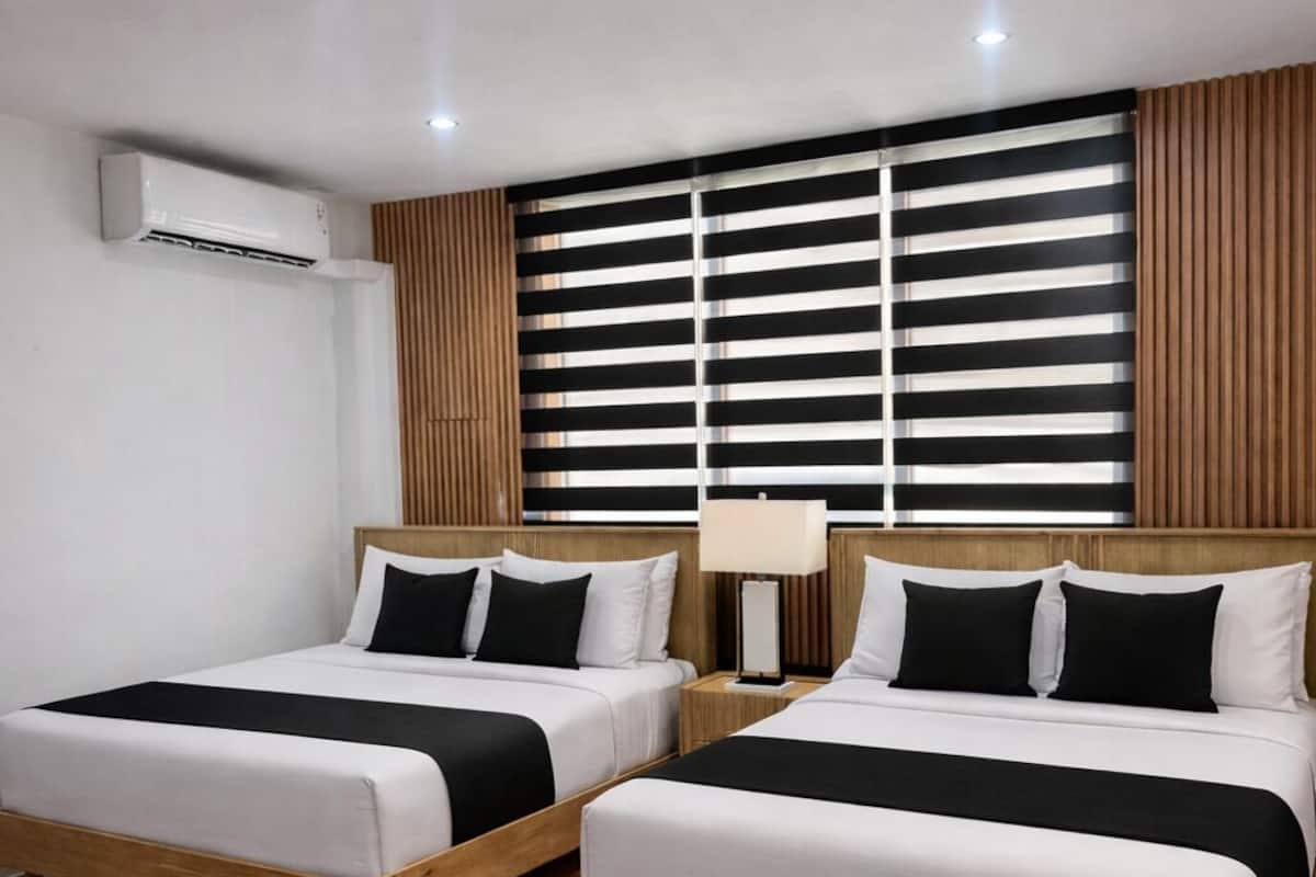 A hotel room with two beds, a wooden floor, a wall-mounted air conditioner, and a window with blinds.