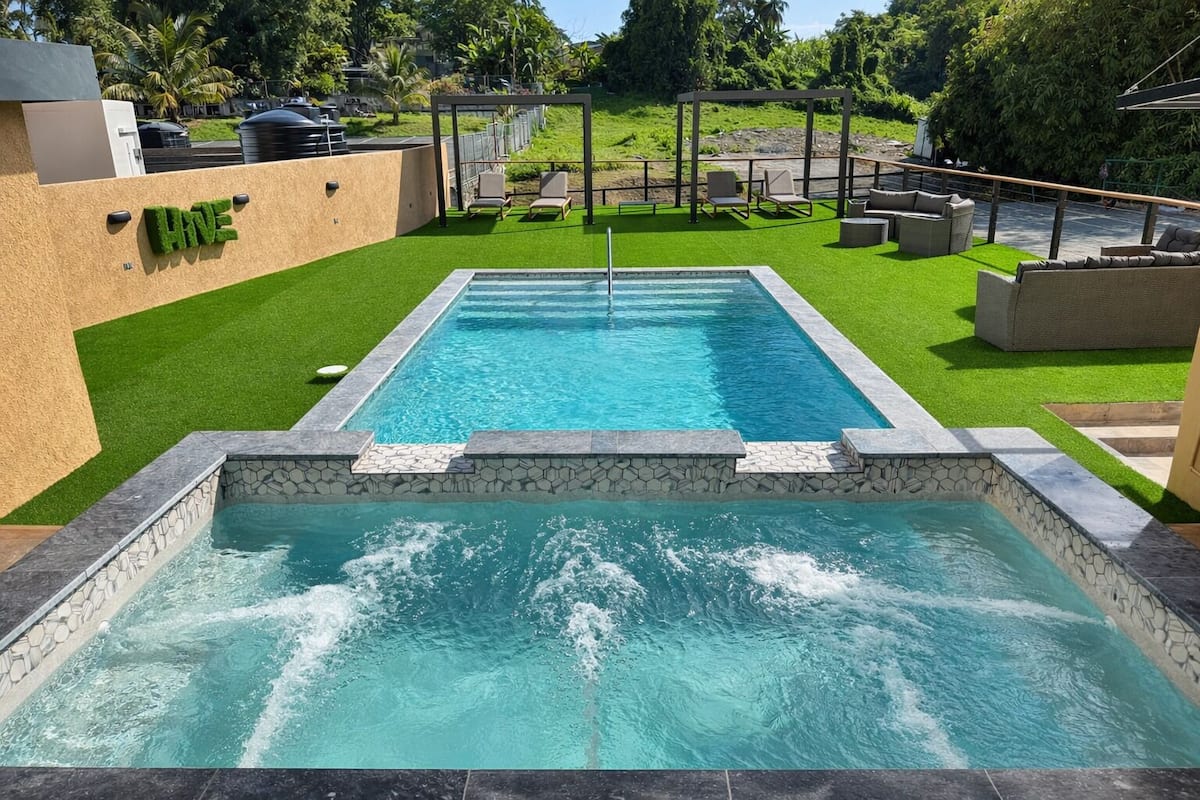 A rectangular swimming pool with a stone border, surrounded by artificial grass and lounge chairs.
