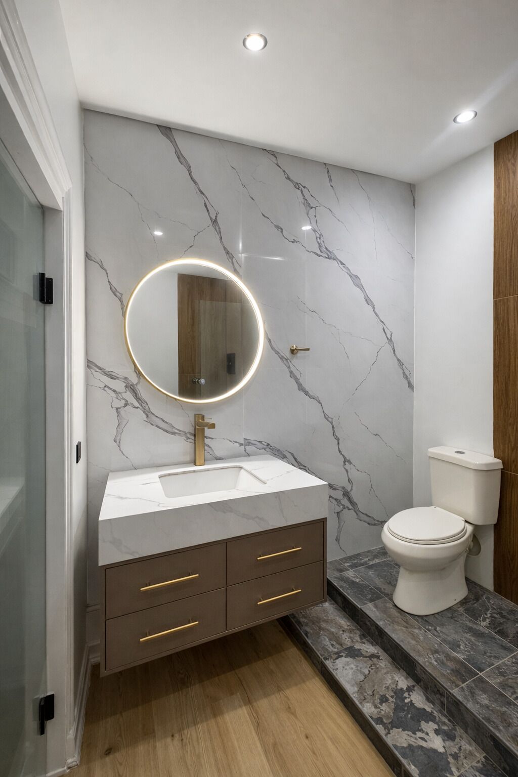 A bathroom with a marble wall, a round mirror with a light, a white sink, a wooden cabinet, and a toilet.
