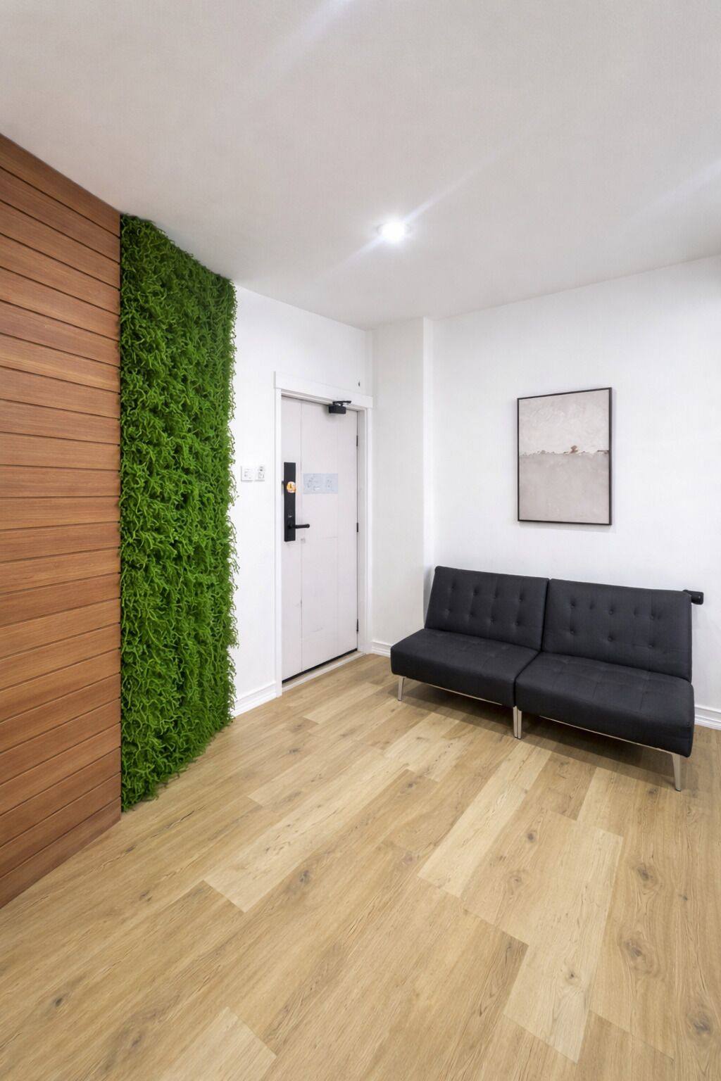A room with a wooden panel wall, a green vertical garden, a black sofa, and a white door.