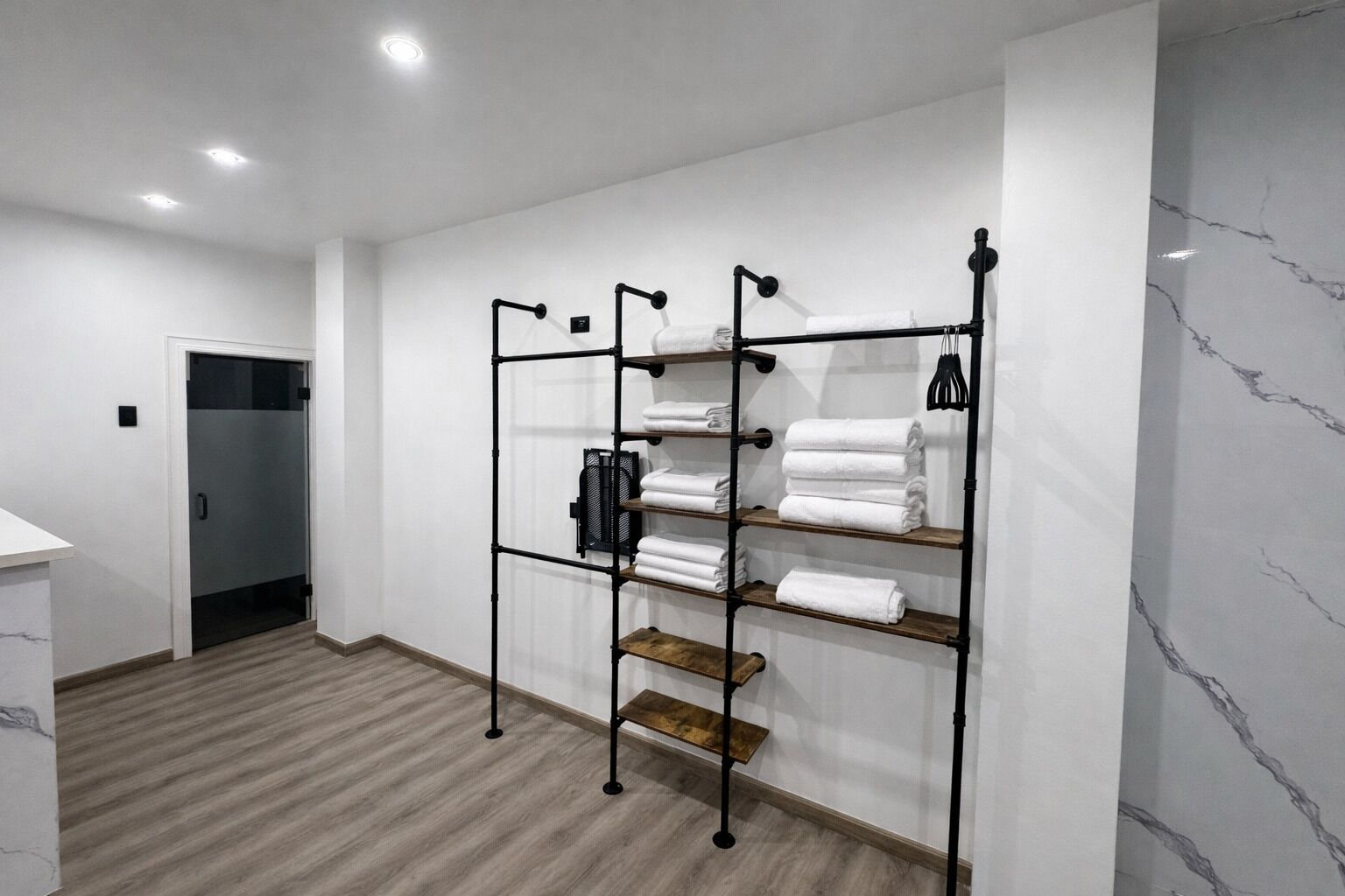 A hotel room with a wooden floor, white walls, a metal rack with folded towels, and a door.