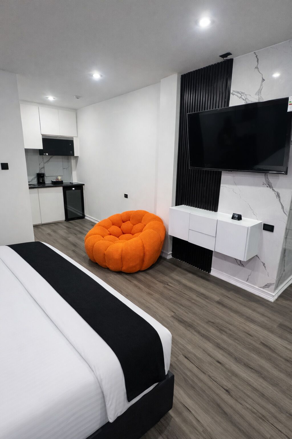 Un dormitorio moderno con cama, televisor, zona de cocina y un original sillón de orejas de conejo naranja.