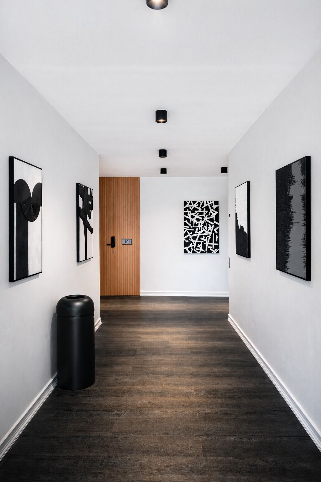 A hallway with dark wooden flooring, white walls, and black artwork. There is a black trash bin on the left and a wooden door at the end.
