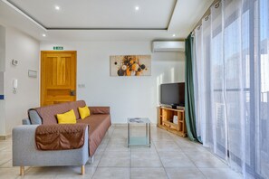 A modern living room with a grey sofa, a small coffee table, a television on a wooden stand, and a large window with sheer curtains. - Charming 2 bedroom flat close to the beach RMUS1-1 (Marsaskala)