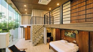 A bedroom with a wooden bed, a small desk, a chair, and a TV. There is a balcony with a chair and a view of greenery.
