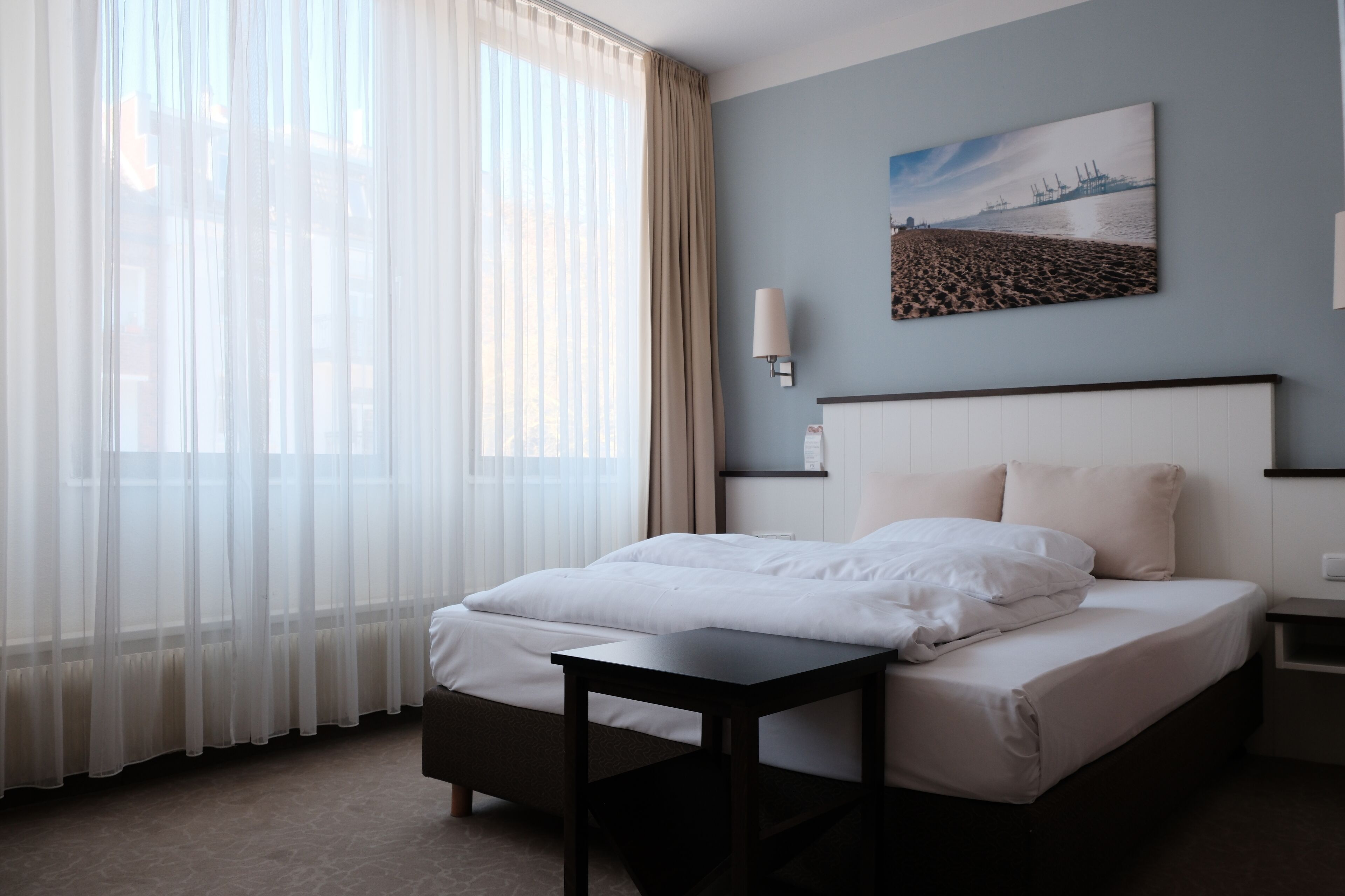 Una habitación de hotel con una cama, una mesita de noche, una lámpara y una ventana con cortinas translúcidas.