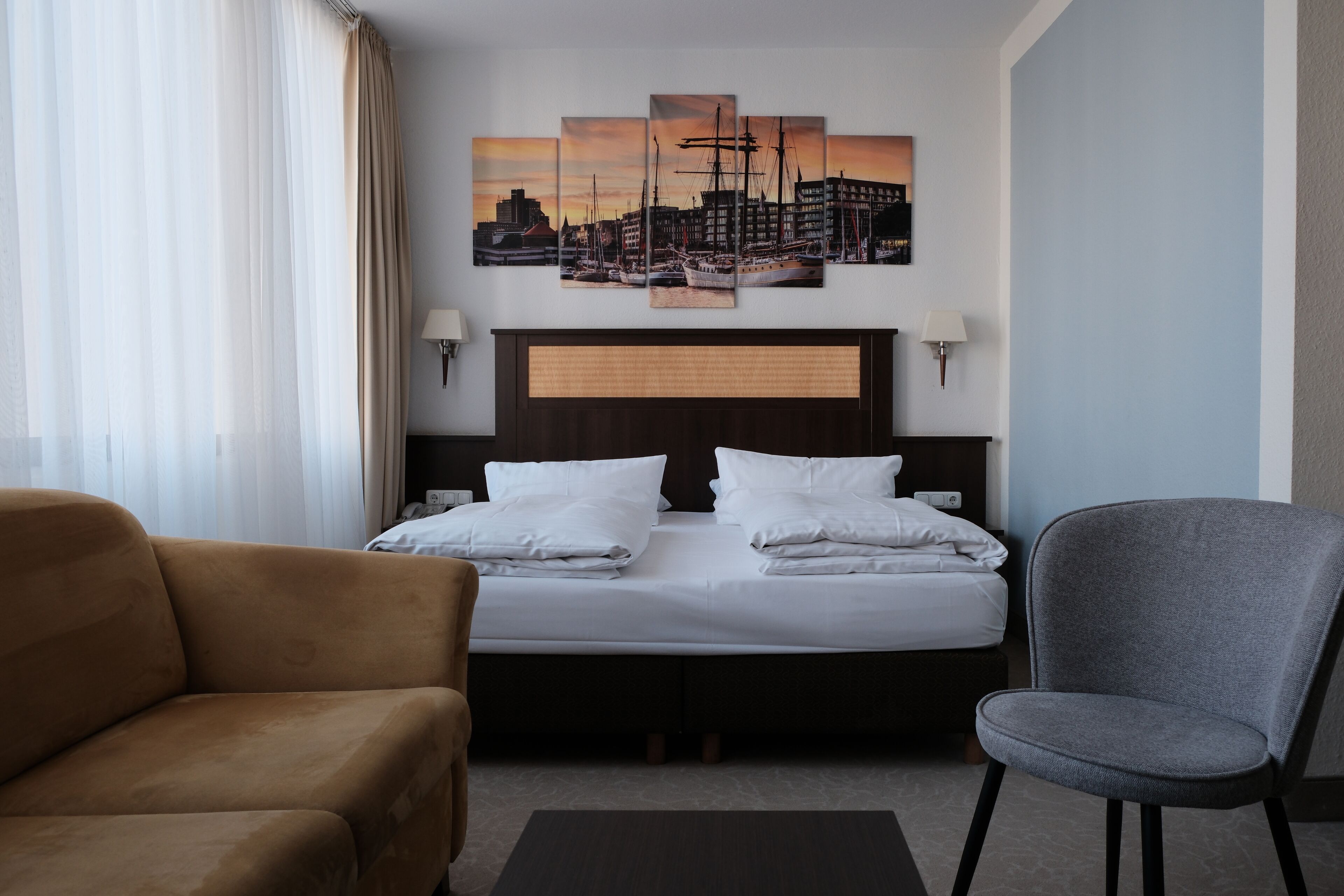 Une chambre d’hôtel avec un lit, un fauteuil marron, un fauteuil gris et un tableau représentant un paysage urbain accroché au mur.