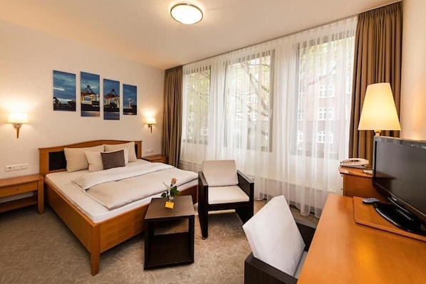 Una habitación de hotel con una cama, un escritorio con televisor, una silla y una mesita con una flor.