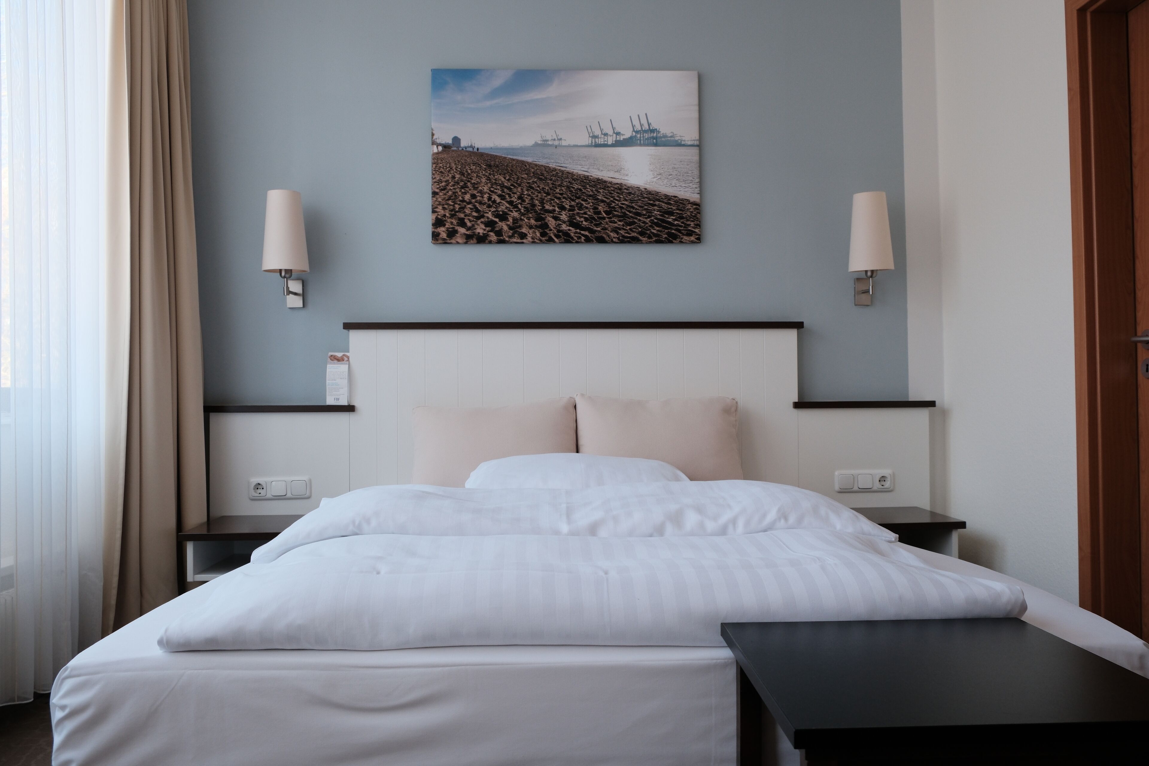 A hotel room with a bed, two bedside tables, a nightstand, a wall-mounted lamp, and a painting of a beach scene.