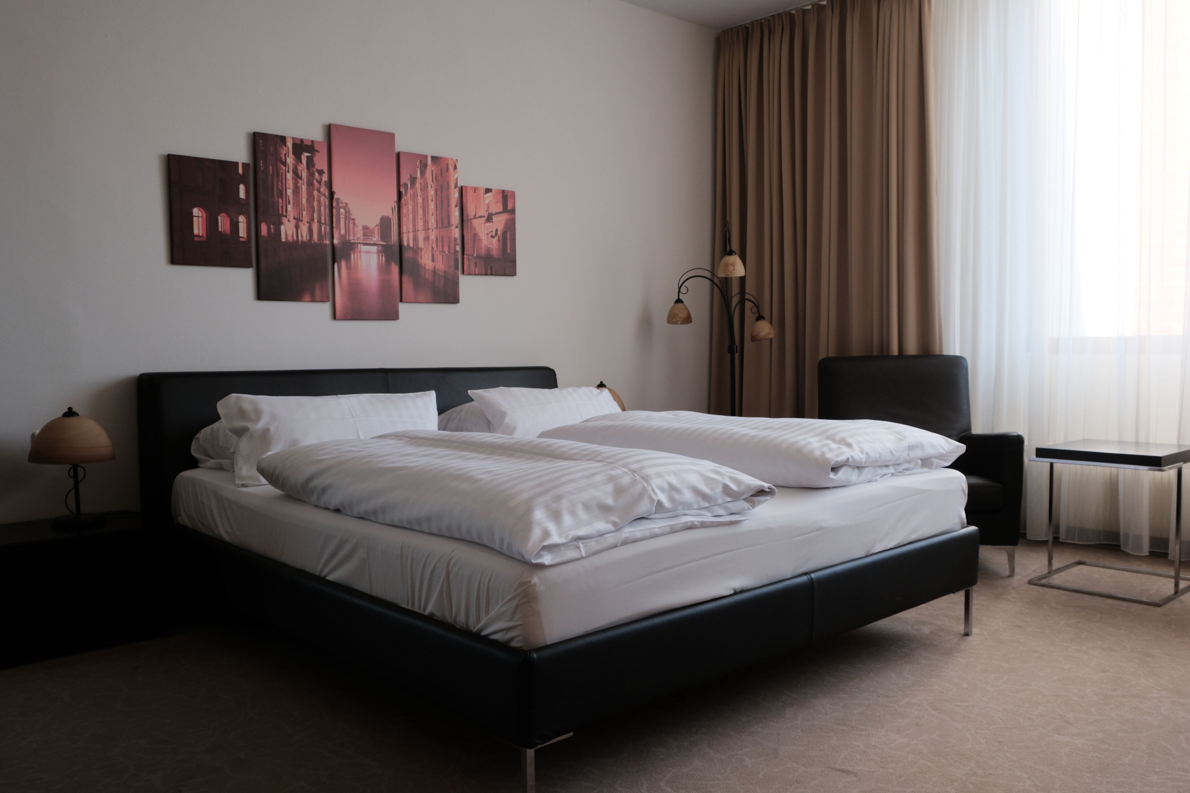 A modern hotel room with a black bed, white linens, a side table with a lamp, a chair, a small table, and a view of the city through sheer curtains.