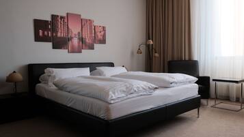 A modern hotel room with a black bed, white linens, a side table with a lamp, a chair, a small table, and a view of the city through sheer curtains.