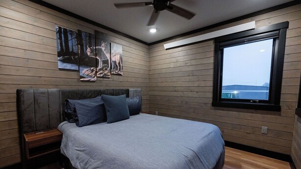 A bedroom with a bed, a nightstand, a ceiling fan, wooden walls, a window with a view, and artwork on the wall. - ATI Unit B: 1  BR, 1  BA Duplex in Sitka, Sleeps 3 (Sitka)