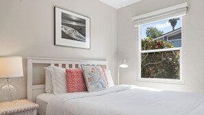 Una camera da letto con un letto bianco, un comodino con una lampada, un quadro appeso al muro e una finestra che si affaccia su alberi e una casa.