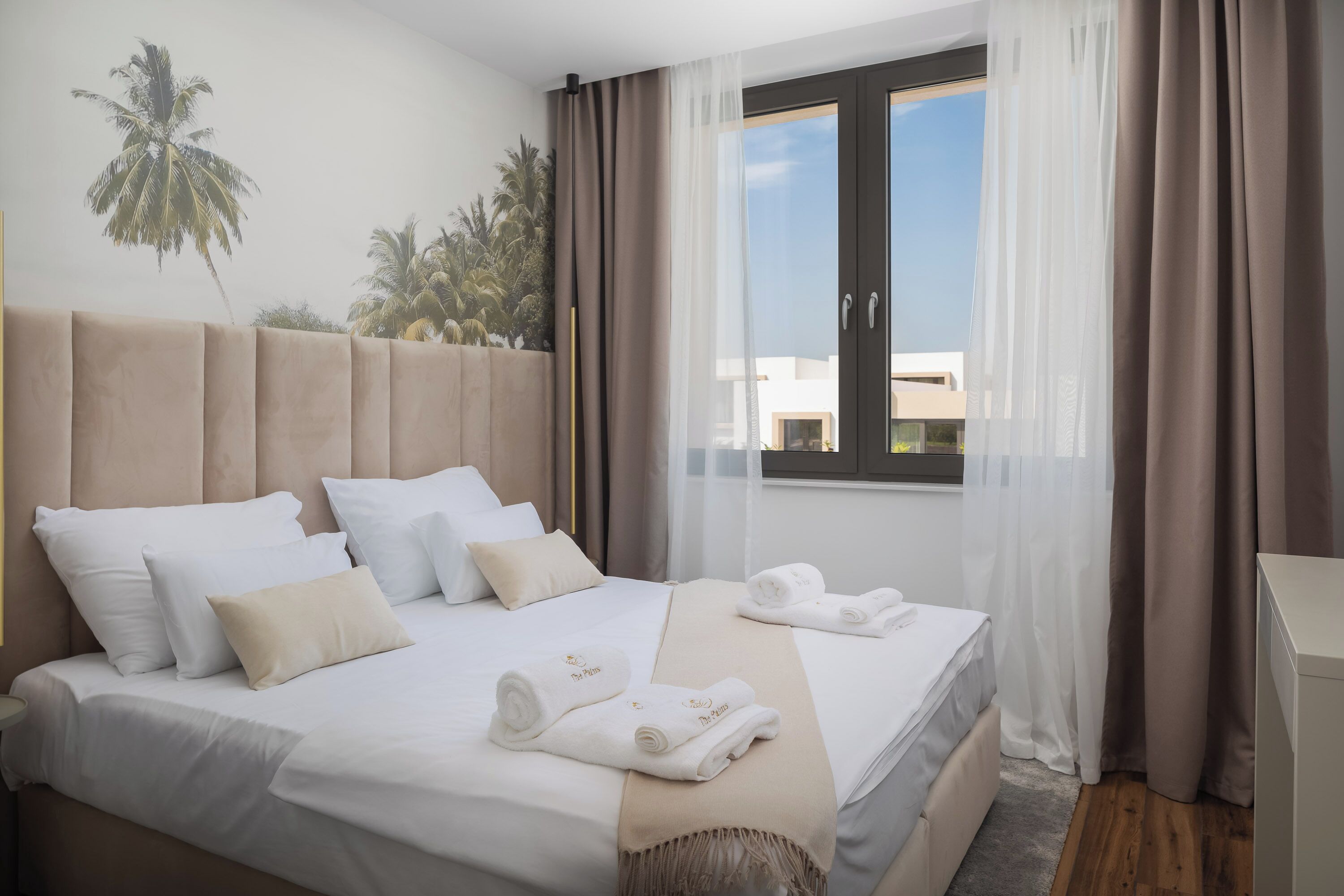 Hotellihuone, jossa on sänky, ikkuna verhoilla ja näkymä palmuille.