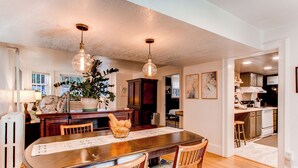 A dining area with a wooden table, chairs, and pendant lights. There is a plant on the table and a view into the kitchen through an open doorway.
