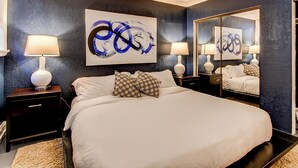 A modern bedroom with a large bed, two bedside tables, a mirror, and abstract wall art.