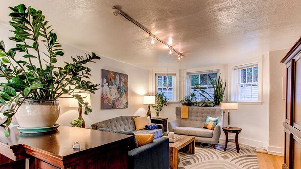 A living room with a sofa, armchairs, a coffee table, and a large potted plant. There are two windows with curtains, a painting on the wall, and a ceiling light fixture.