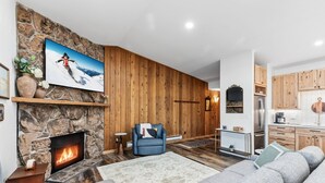 A living room with a stone fireplace, a blue armchair, a sofa, and a kitchen area with wooden cabinets. - Buffalo Mtn Views - Spacious Dillon Retreat (Dillon)