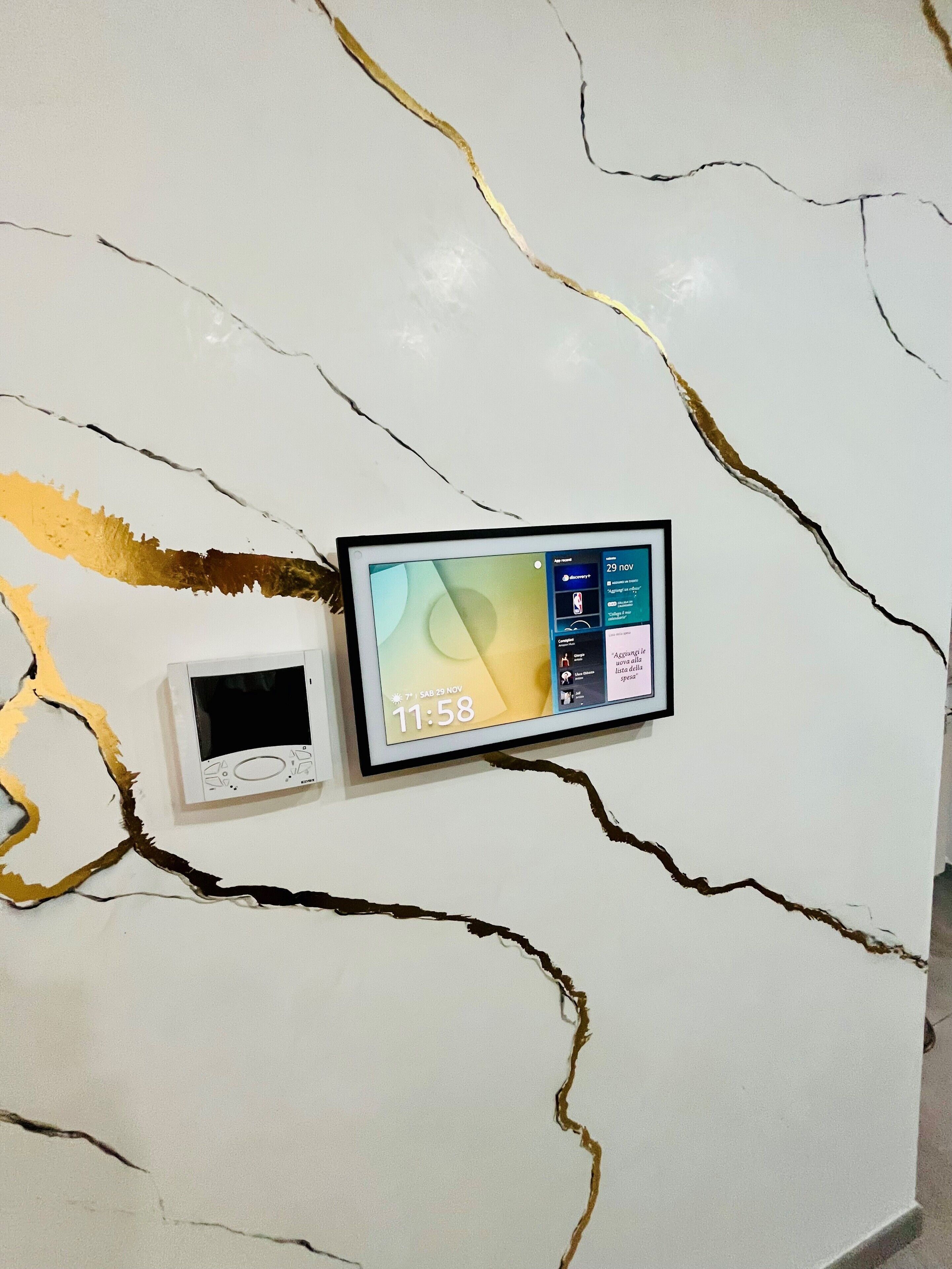 Una pared con diseño de mármol blanco con vetas doradas, un reloj digital enmarcado que muestra la hora, y un dispositivo digital más pequeño debajo.