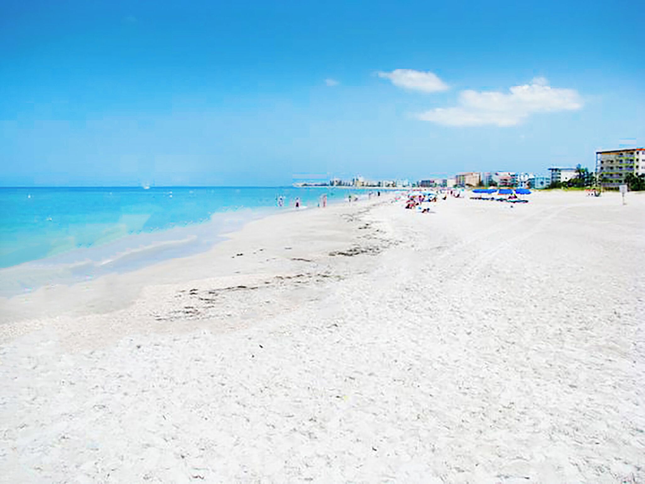 A wide sandy beach with clear blue water, a few people, and buildings in the background.