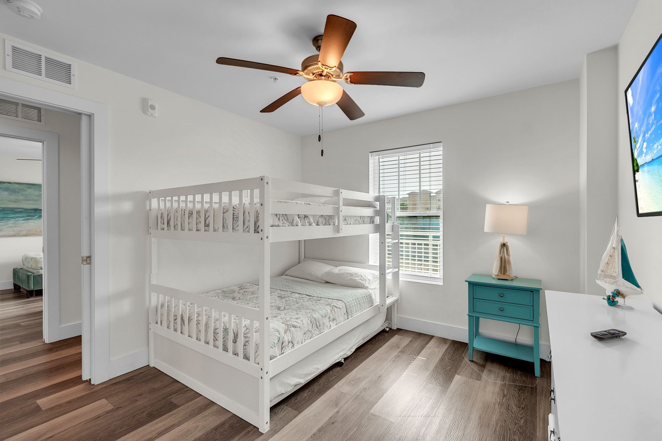 Una cama litera infantil en una habitación con ventilador de techo, ventana con persianas, mesita de noche color turquesa y un cuadro en la pared.