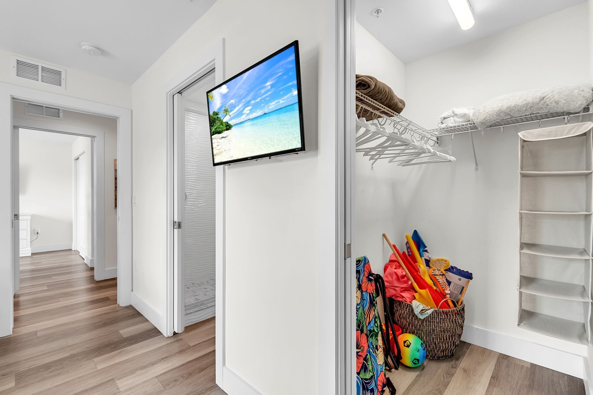 Un couloir avec un téléviseur diffusant une scène de plage, un panier en osier contenant des objets colorés et une étagère blanche.