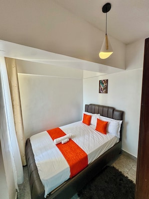 Un lit simple avec une tête de lit, du linge de lit blanc et des touches de couleur orange, dans une pièce dotée d’un luminaire au plafond et d’un tableau au mur.