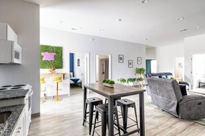 Eine moderne Küche mit integriertem Mikrowellenherd, ein Essbereich mit Holztisch und Barhocker sowie ein Wohnbereich mit grauem Sofa und grauem Sessel.