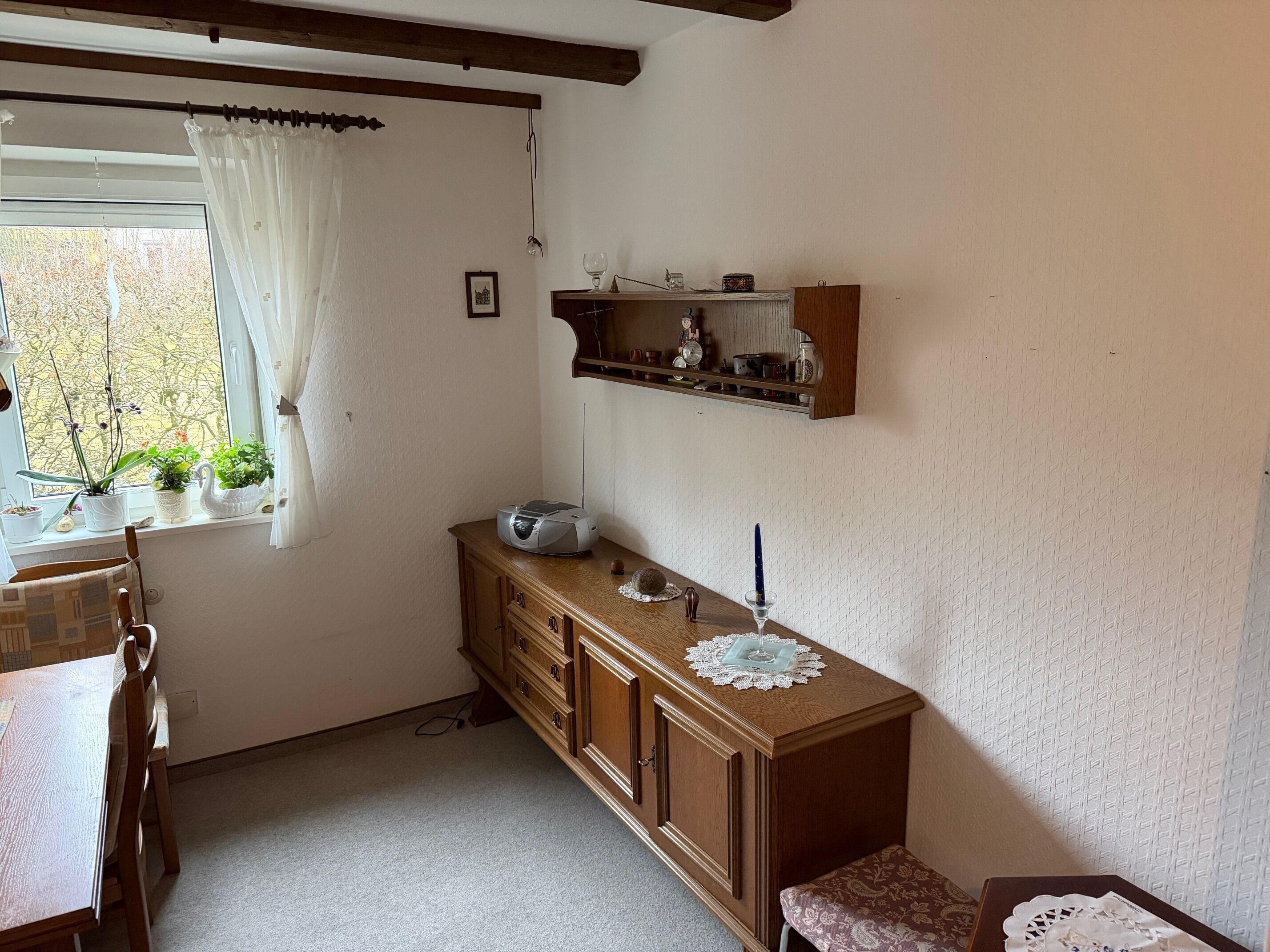 A small, cozy kitchen with a wooden cabinet, a window with white curtains, and a shelf above the cabinet.