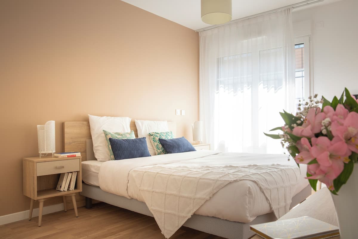 Une chambre à coucher avec un lit, des tables de chevet, une commode, une fenêtre avec des rideaux légers et un vase rempli de fleurs.
