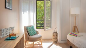 Une chambre à coucher comprenant un lit, une chaise, un bureau avec une lampe, une fenêtre avec des rideaux et une vue sur la verdure à l’extérieur.