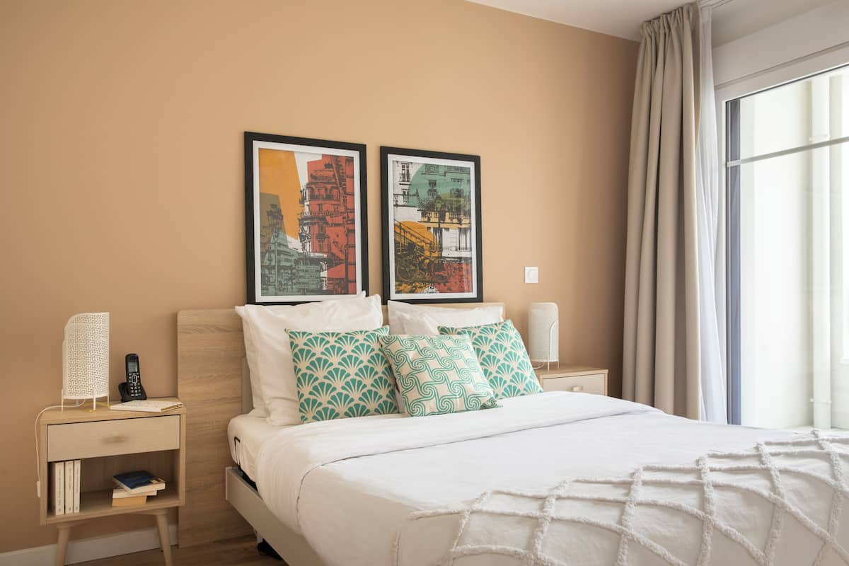 Une chambre d’hôtel avec un lit, deux tables de chevet, une commode, une lampe, un tableau encadré et une fenêtre avec des rideaux.