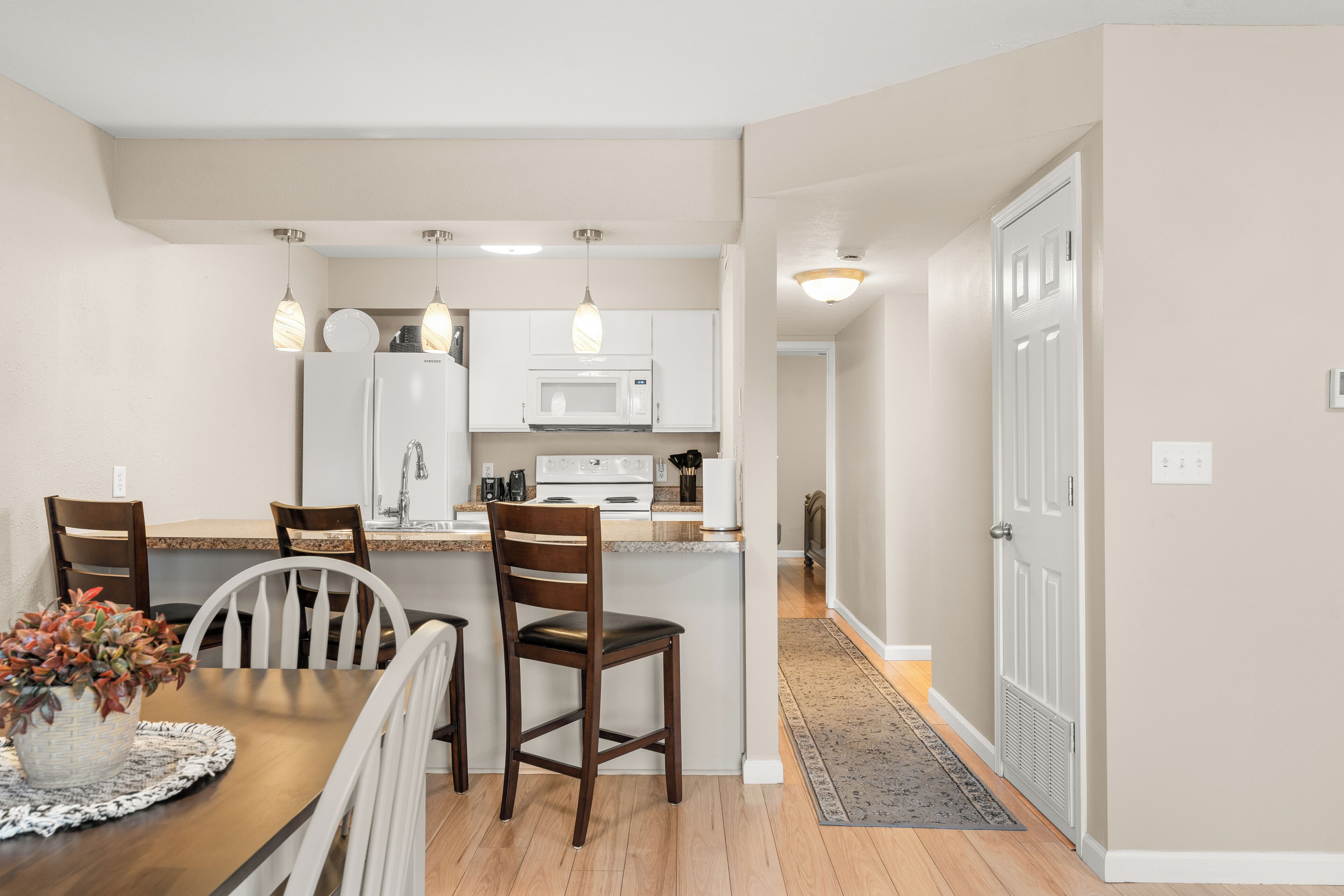 A modern kitchen with white cabinets, a microwave, and a refrigerator. There is a dining area with a wooden table and chairs.