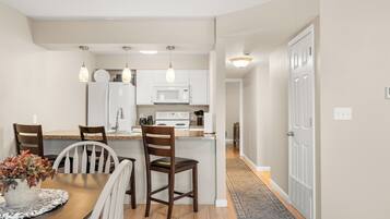 A modern kitchen with white cabinets, a microwave, and a refrigerator. There is a dining area with a wooden table and chairs.