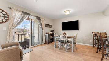 A spacious living area with a dining table and chairs, a sofa, a television mounted on the wall, and a sliding glass door leading to an outdoor balcony.