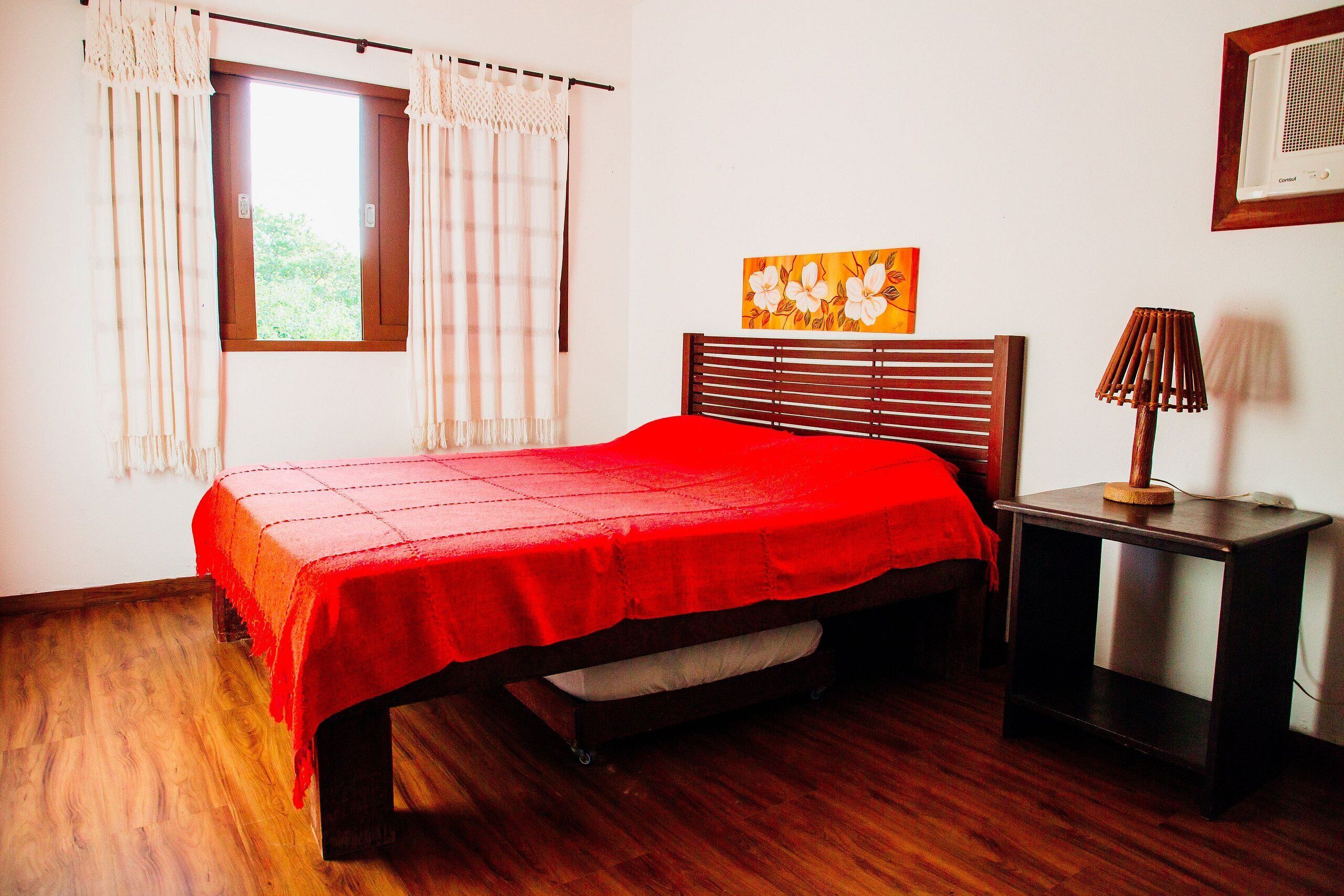 A bedroom with a wooden bed, a red bedspread, a nightstand with a lamp, and a window with curtains.