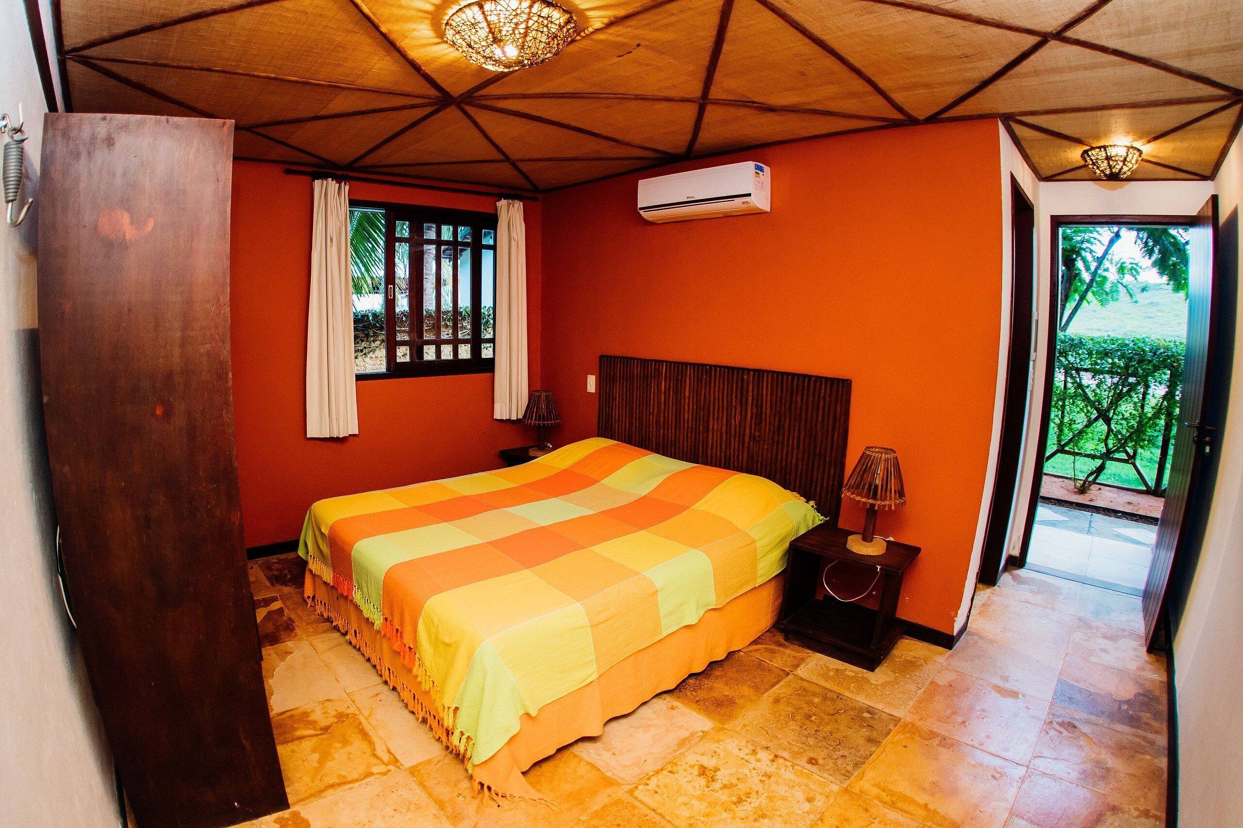A bedroom with a bed, an air conditioner, a nightstand, a lamp, and a view of greenery outside.